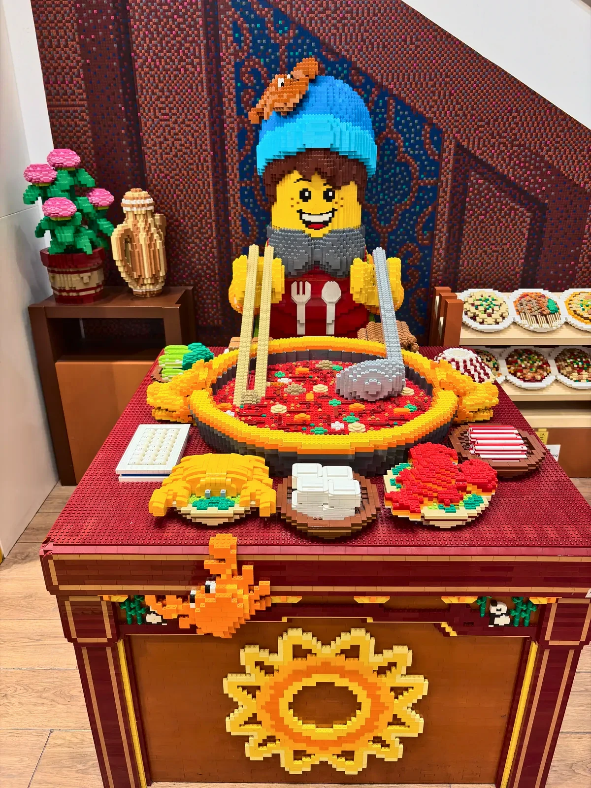 Brick-built figure in a beanie and bib at a hotpot table of miniature brick dishes